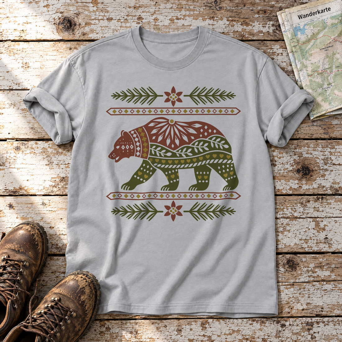 Bär im Alpenornament – T-Shirt Unisex in Hellgrau, flach liegend auf alten Holzbrettern mit Wanderschuhen und Wanderkarte.