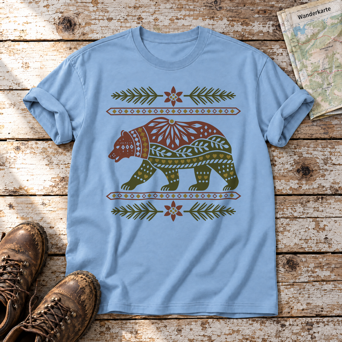 Bär im Alpenornament – T-Shirt Unisex in Himmelblau, flach liegend auf alten Holzbrettern mit Wanderschuhen und Wanderkarte.