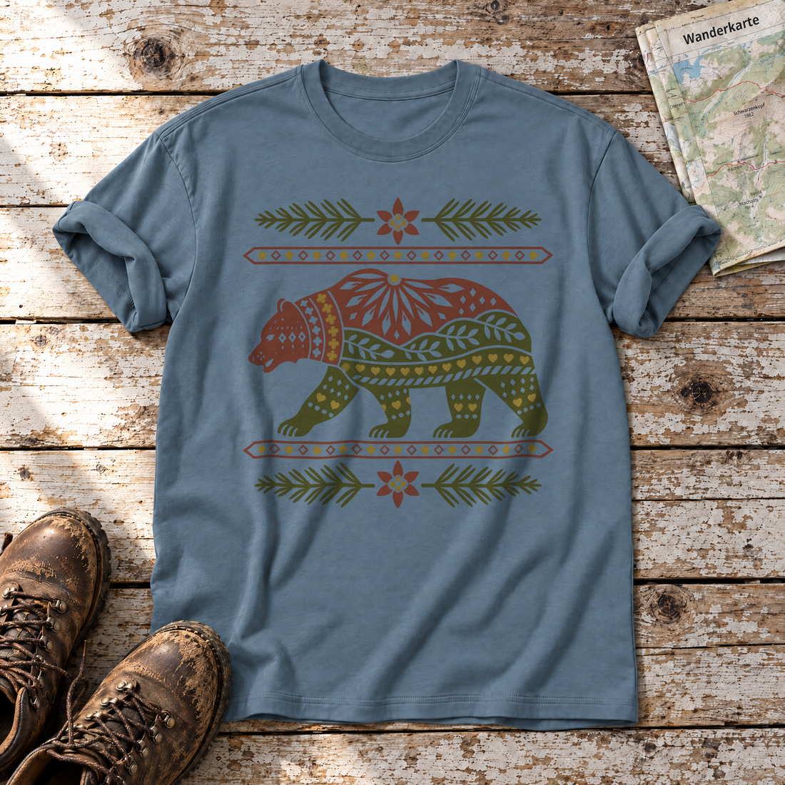 Bär im Alpenornament – T-Shirt Unisex in Steinblau, flach liegend auf alten Holzbrettern mit Wanderschuhen und Wanderkarte.