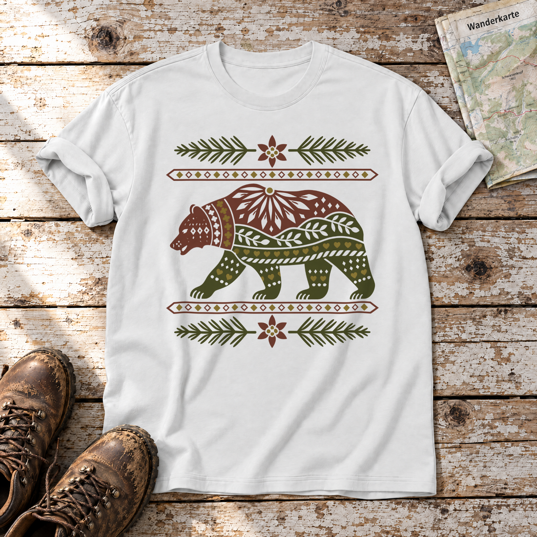 Bär im Alpenornament – T-Shirt Unisex in Weiß, flach liegend auf alten Holzbrettern mit Wanderschuhen und Wanderkarte.