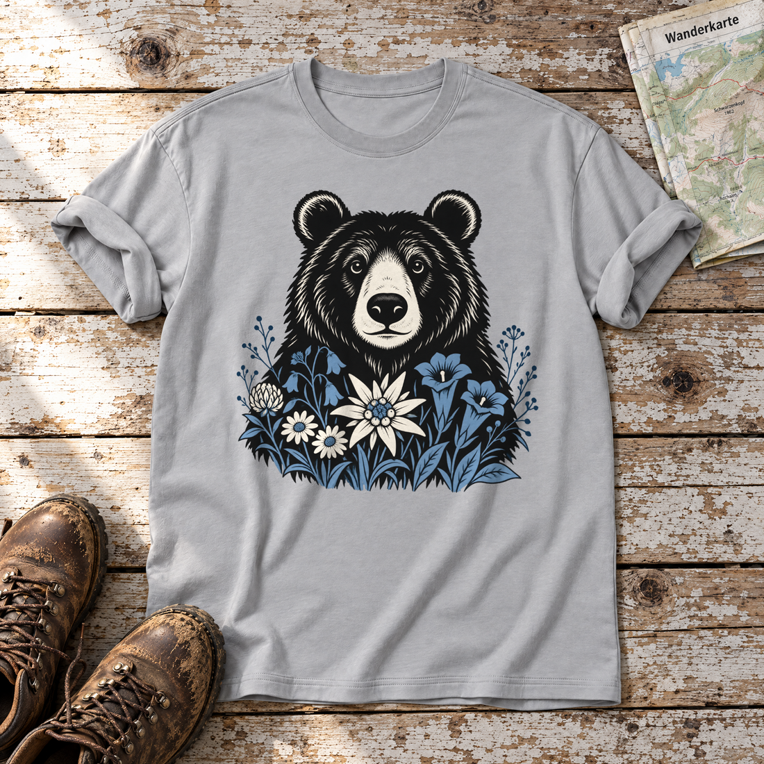 Bär in der Alpenwiese – T-Shirt Unisex in Hellgrau, flach liegend auf alten Holzbrettern mit Wanderschuhen und Wanderkarte.