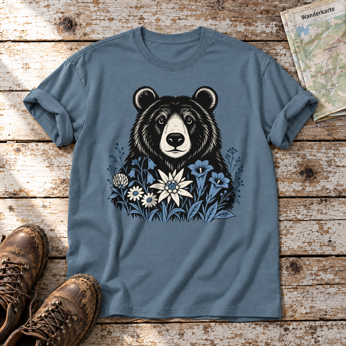 Bär in der Alpenwiese – T-Shirt Unisex in Steinblau, flach liegend auf alten Holzbrettern mit Wanderschuhen und Wanderkarte.