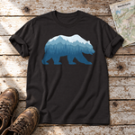 Bär im Bergwald – T-Shirt Unisex in Schwarz, flach liegend auf alten Holzbrettern mit Wanderschuhen und Wanderkarte.