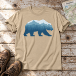 Bär im Bergwald – T-Shirt Unisex in Sand, flach liegend auf alten Holzbrettern mit Wanderschuhen und Wanderkarte.