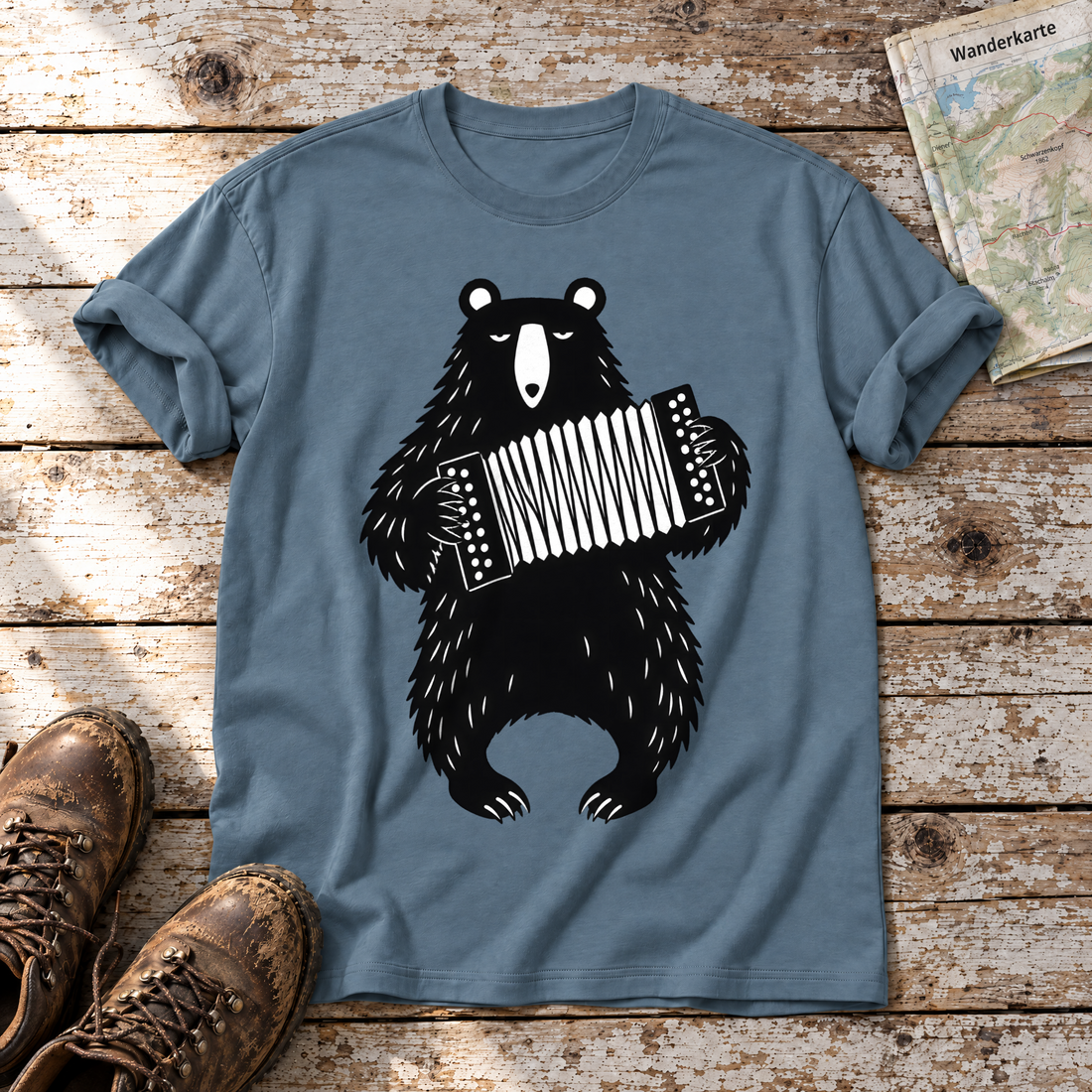 Bär mit Steirischer – T-Shirt Unisex in Steinblau, flach liegend auf alten Holzbrettern mit Wanderschuhen und Wanderkarte.