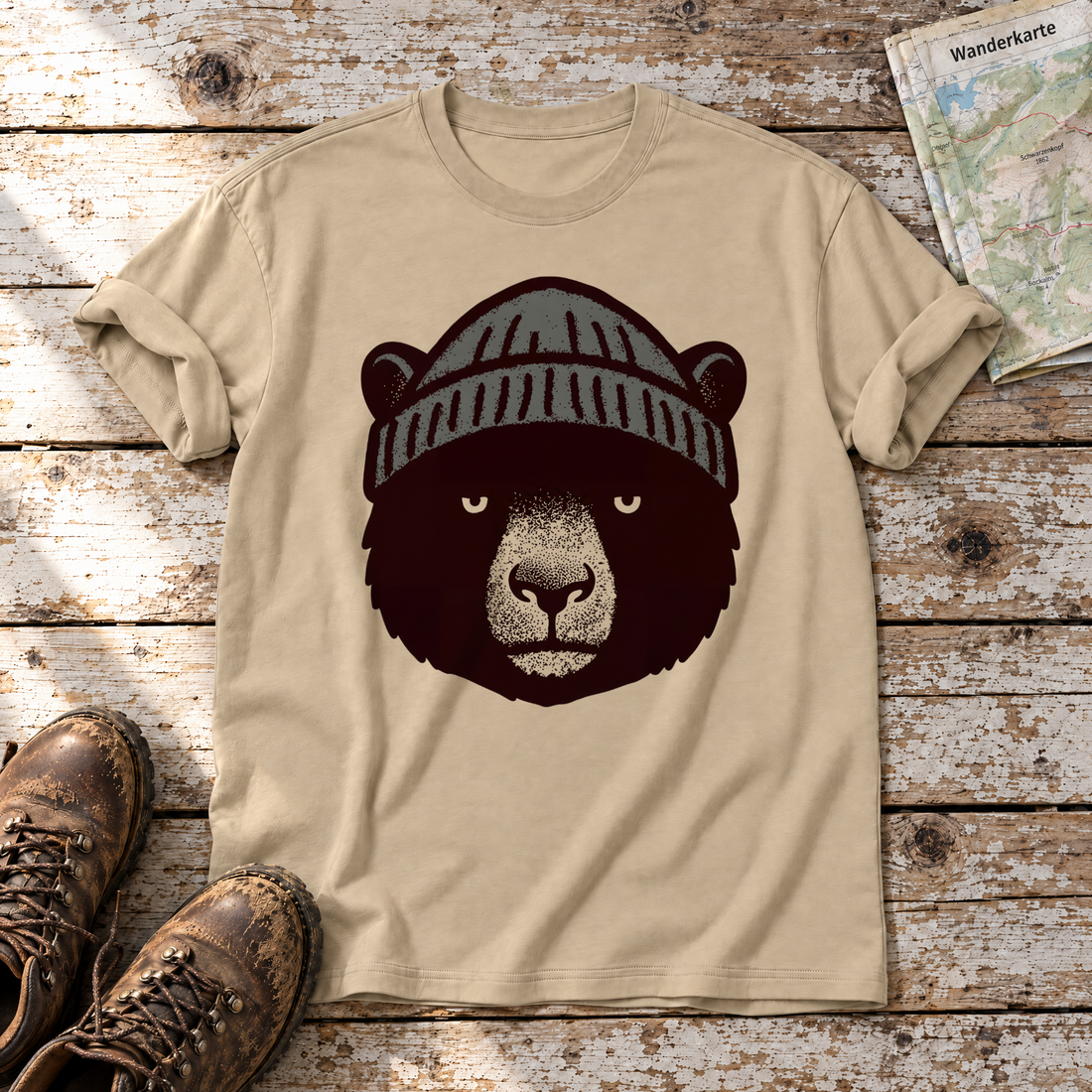 Bär mit Strickmütze – T-Shirt Unisex in Sand, flach liegend auf alten Holzbrettern mit Wanderschuhen und Wanderkarte.