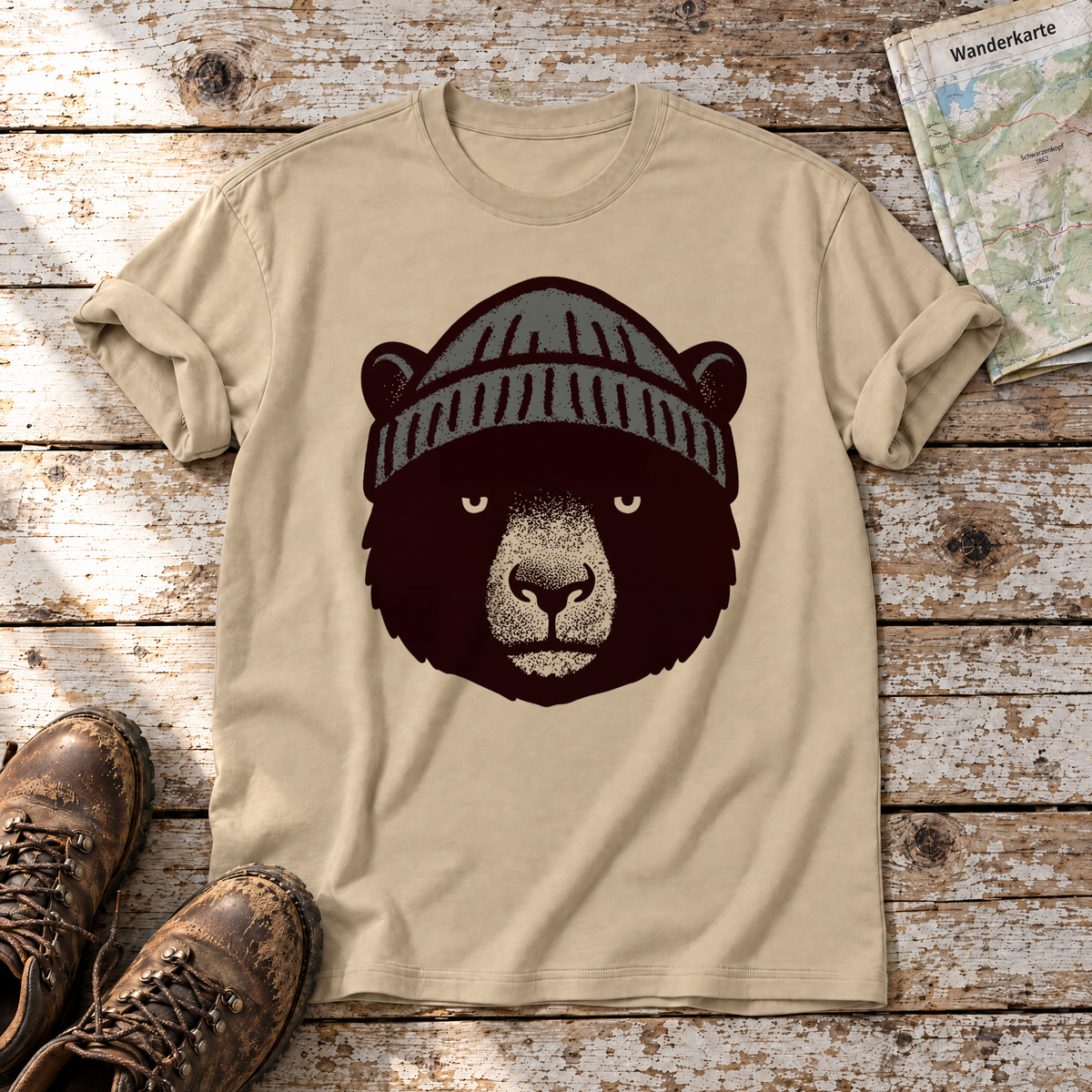 Bär mit Strickmütze – T-Shirt Unisex in Sand, flach liegend auf alten Holzbrettern mit Wanderschuhen und Wanderkarte.