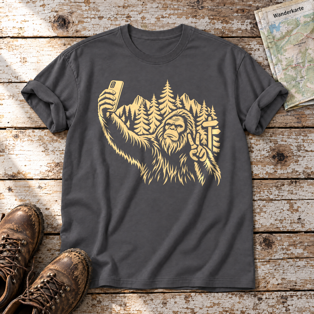 Bigfoot-Selfie im Bergwald – T-Shirt Unisex in Dunkelgrau, flach liegend auf alten Holzbrettern mit Wanderschuhen und Wanderkarte.