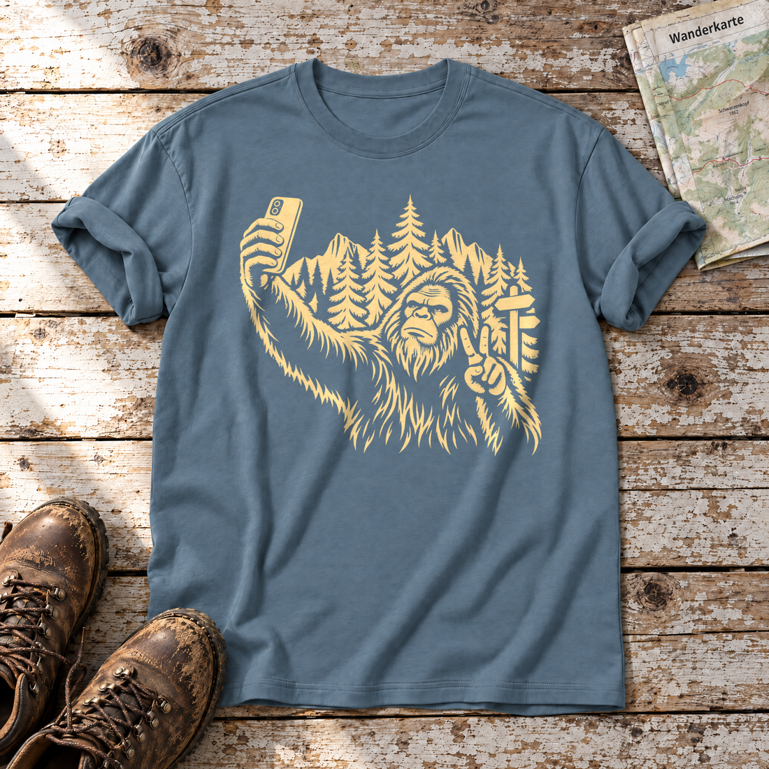 Bigfoot-Selfie im Bergwald – T-Shirt Unisex in Steinblau, flach liegend auf alten Holzbrettern mit Wanderschuhen und Wanderkarte.