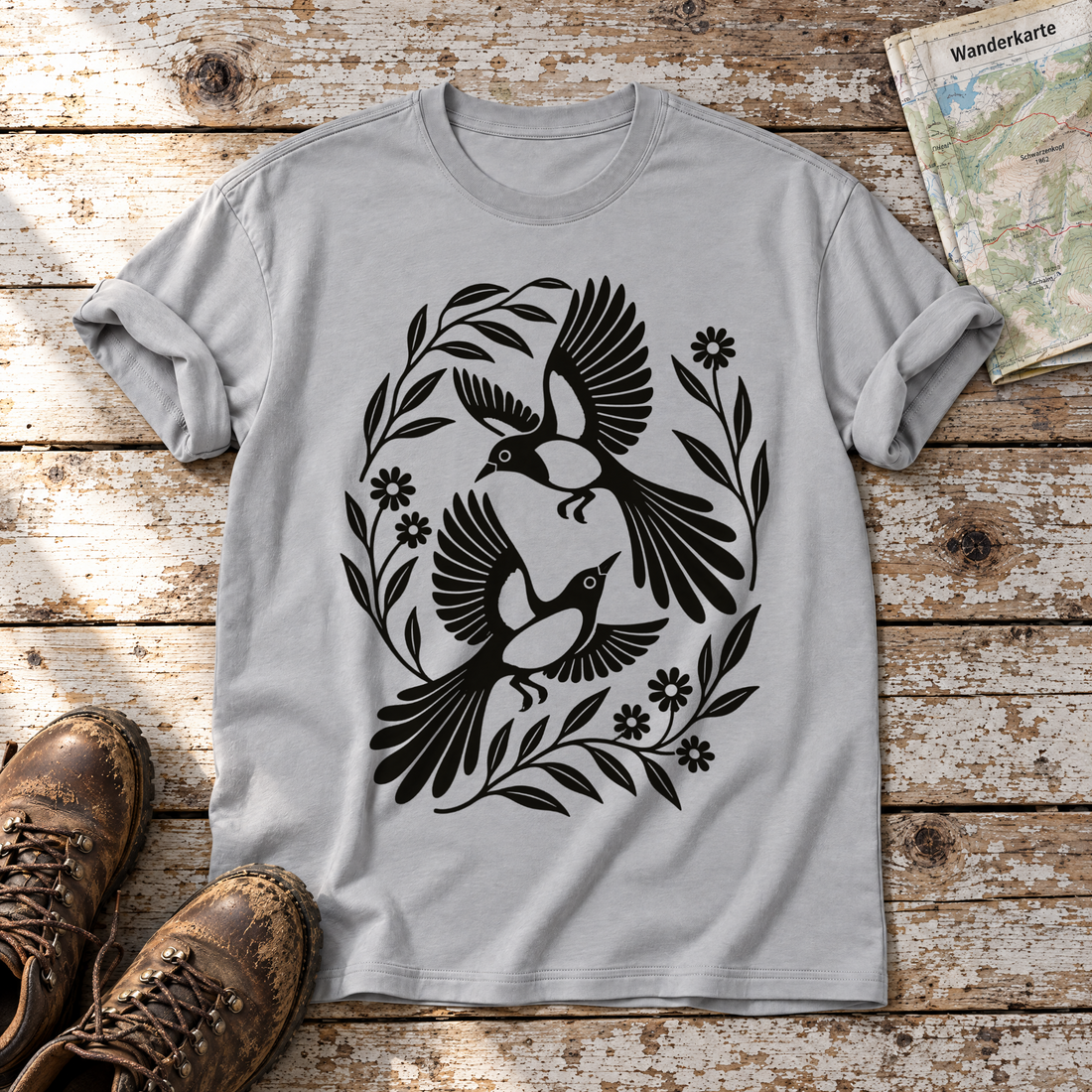Elsternpaar im Scherenschnitt – T-Shirt Unisex in Hellgrau, flach liegend auf alten Holzbrettern mit Wanderschuhen und Wanderkarte.
