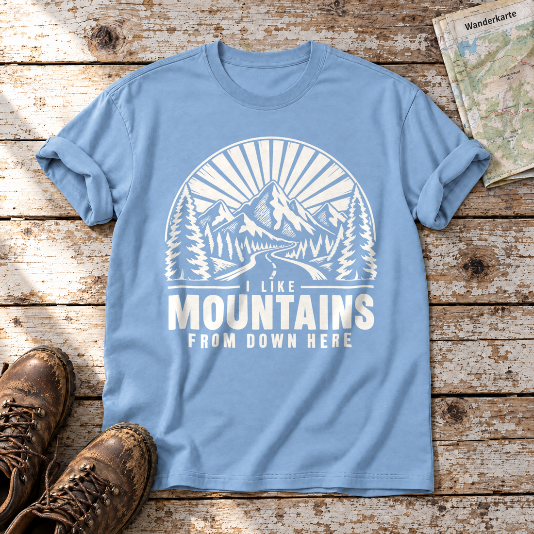 I like mountains from down here – T-Shirt Unisex in Himmelblau, flach liegend auf alten Holzbrettern mit Wanderschuhen und Wanderkarte.