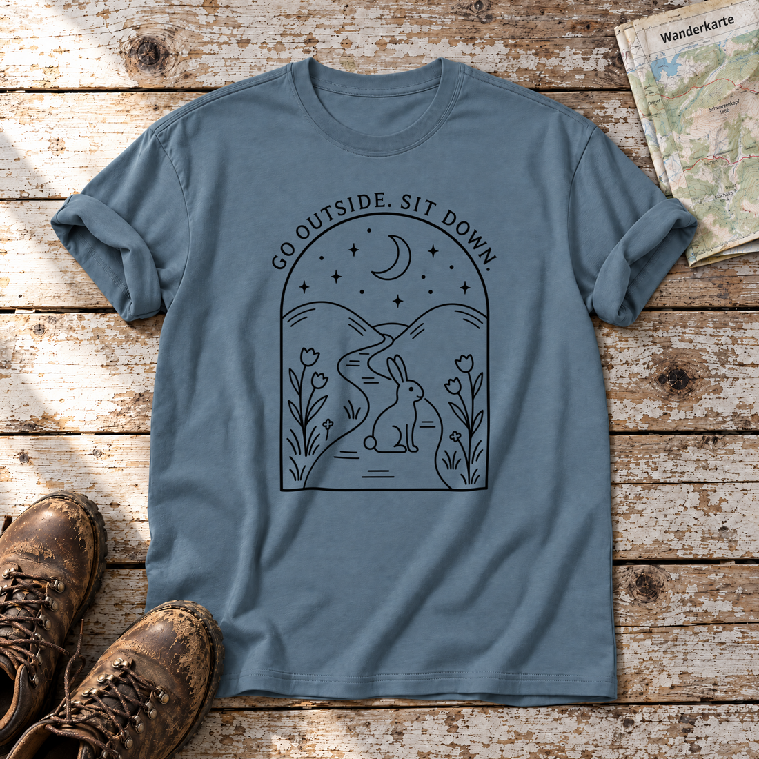 Go outside. Sit down. – T-Shirt Unisex in Steinblau, flach liegend auf alten Holzbrettern mit Wanderschuhen und Wanderkarte.