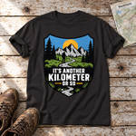 Another Kilometer or So – T-Shirt Unisex in Schwarz, flach liegend auf alten Holzbrettern mit Wanderschuhen und Wanderkarte.