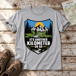 Another Kilometer or So – T-Shirt Unisex in Hellgrau, flach liegend auf alten Holzbrettern mit Wanderschuhen und Wanderkarte.