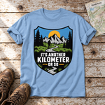 Another Kilometer or So – T-Shirt Unisex in Himmelblau, flach liegend auf alten Holzbrettern mit Wanderschuhen und Wanderkarte.