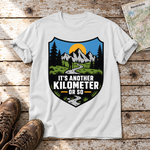Another Kilometer or So – T-Shirt Unisex in Weiß, flach liegend auf alten Holzbrettern mit Wanderschuhen und Wanderkarte.