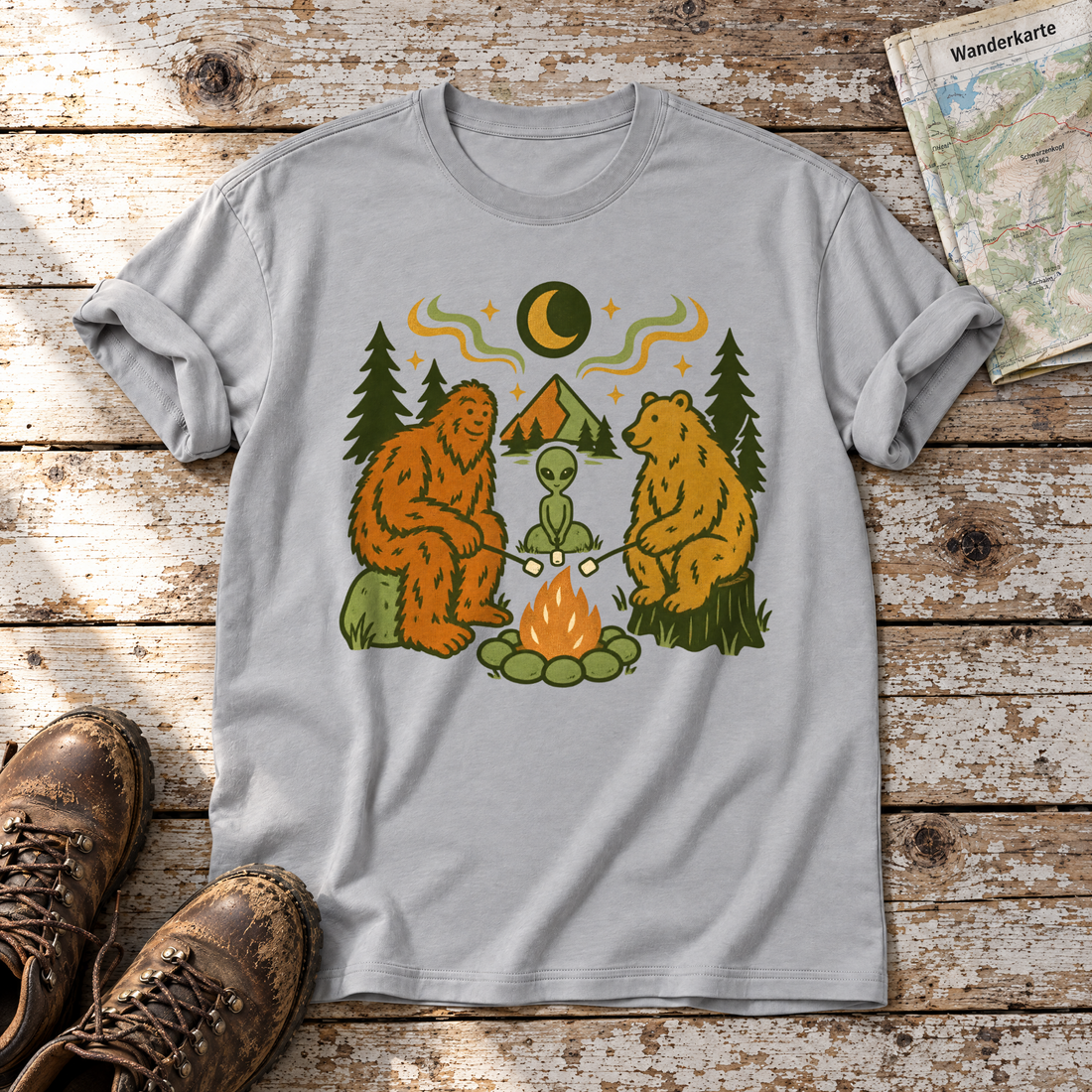 Bigfoot, Alien und Bär am Lagerfeuer – T-Shirt Unisex in Hellgrau, flach liegend auf alten Holzbrettern mit Wanderschuhen und Wanderkarte.