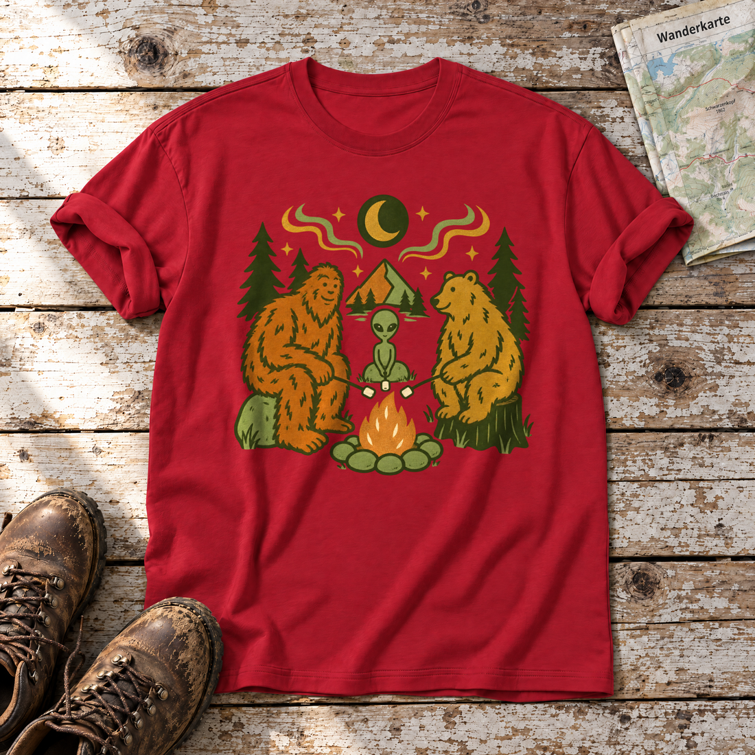 Bigfoot, Alien und Bär am Lagerfeuer – T-Shirt Unisex in Rot, flach liegend auf alten Holzbrettern mit Wanderschuhen und Wanderkarte.