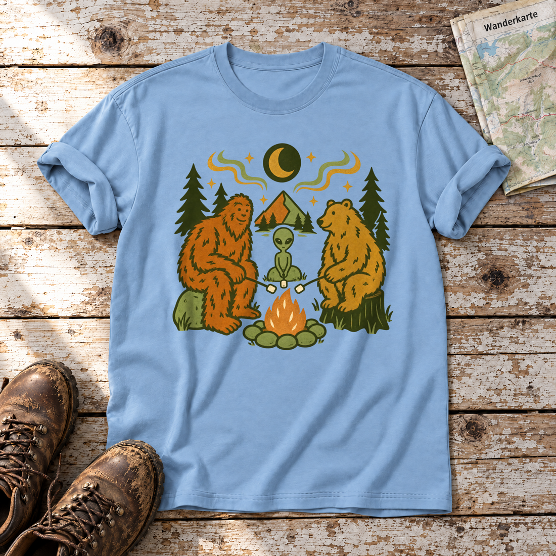 Bigfoot, Alien und Bär am Lagerfeuer – T-Shirt Unisex in Himmelblau, flach liegend auf alten Holzbrettern mit Wanderschuhen und Wanderkarte.