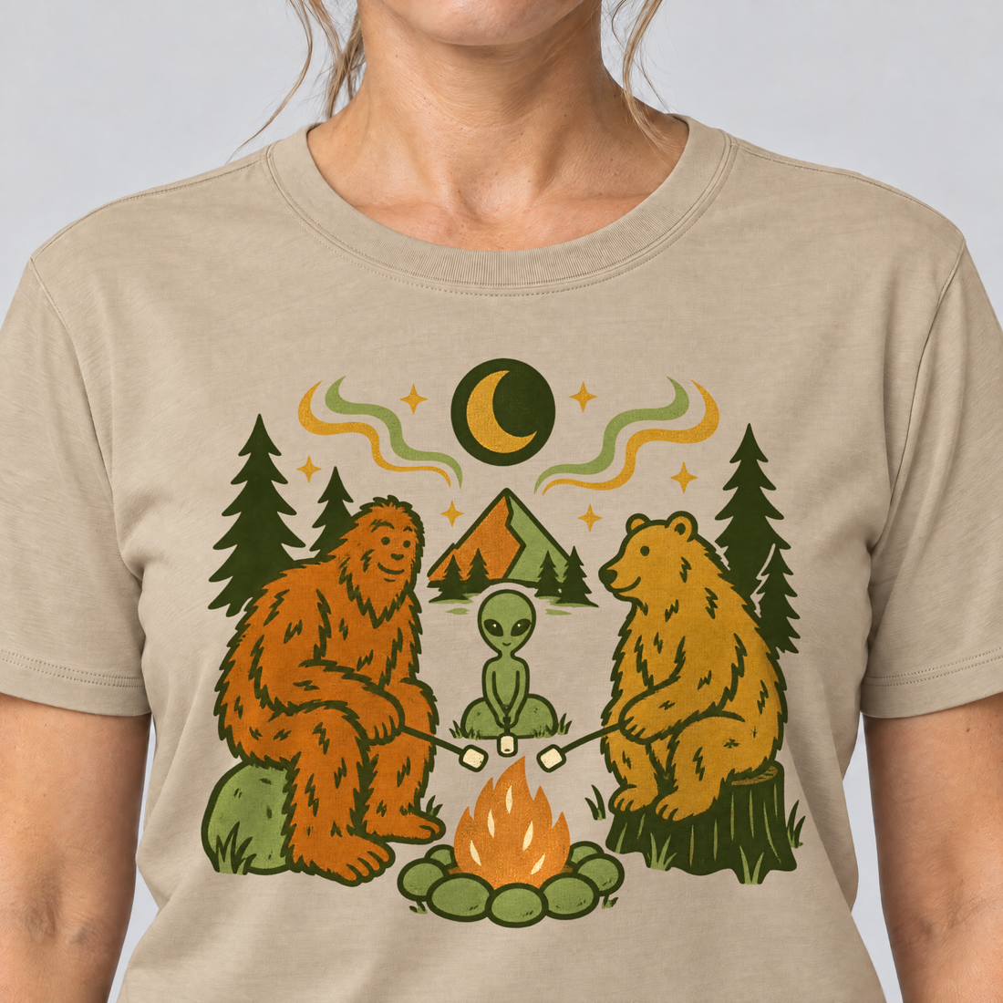 Nahaufnahme des Drucks: Bigfoot, Alien und Bär am Lagerfeuer – T-Shirt Unisex in Sand, von einer Frau getragen