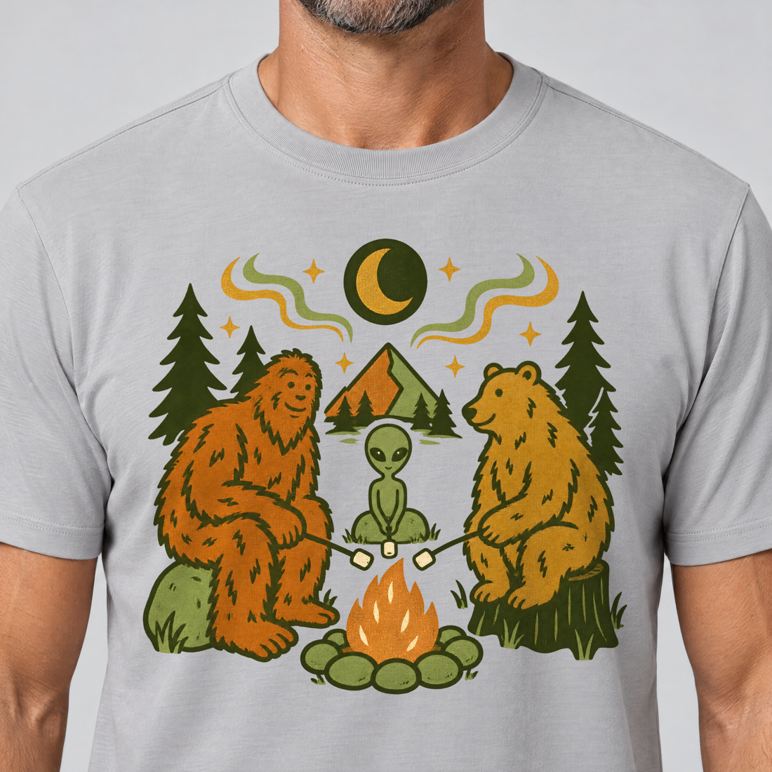Nahaufnahme des Drucks: Bigfoot, Alien und Bär am Lagerfeuer – T-Shirt Unisex in Hellgrau, von einem Mann getragen
