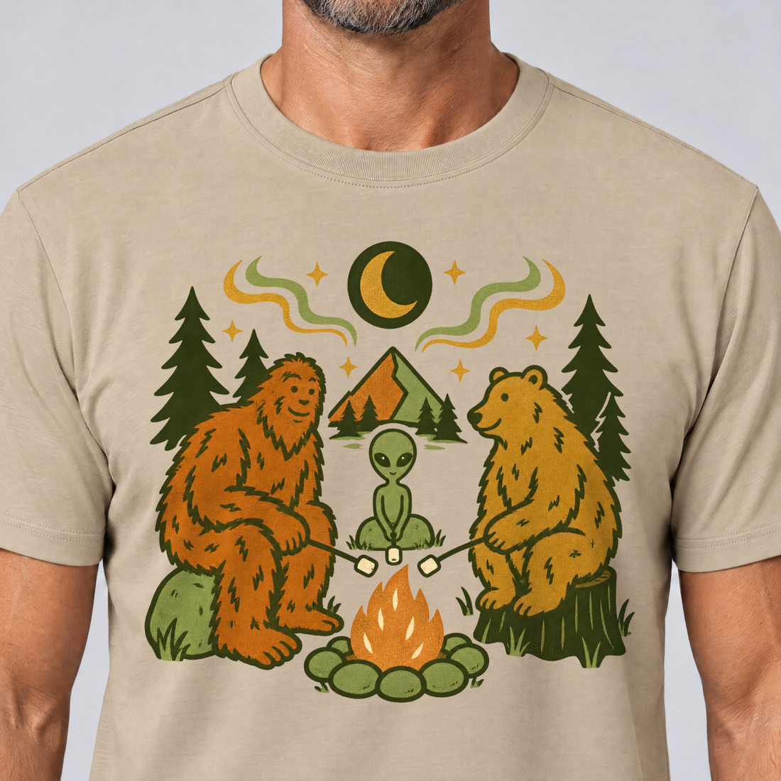 Nahaufnahme des Drucks: Bigfoot, Alien und Bär am Lagerfeuer – T-Shirt Unisex in Sand, von einem Mann getragen