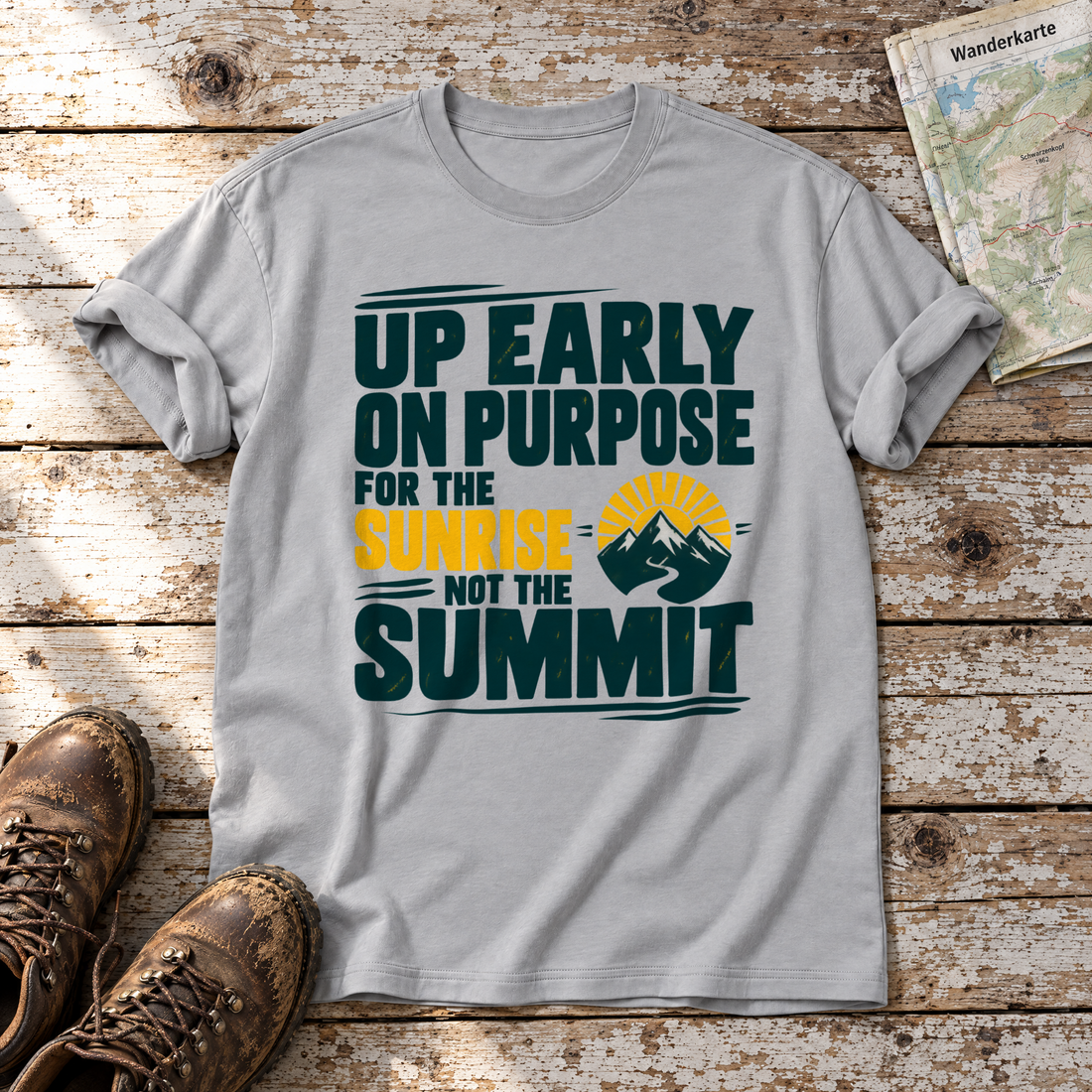 For the Sunrise, Not the Summit – T-Shirt Unisex in Hellgrau, flach liegend auf alten Holzbrettern mit Wanderschuhen und Wanderkarte.