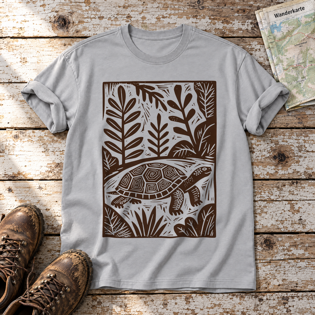 Schildkröte im Linolschnitt – T-Shirt Unisex in Hellgrau, flach liegend auf alten Holzbrettern mit Wanderschuhen und Wanderkarte.