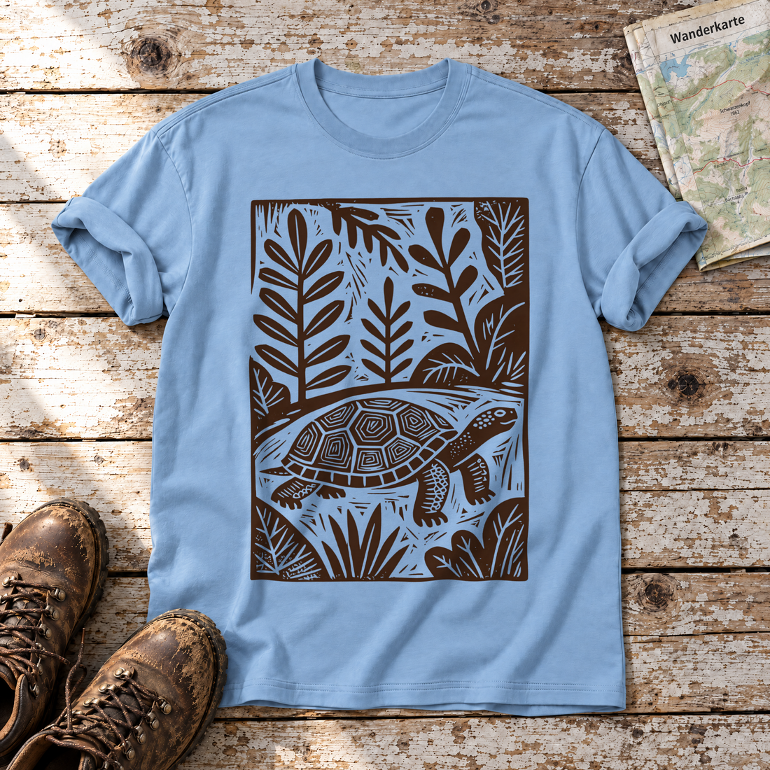 Schildkröte im Linolschnitt – T-Shirt Unisex in Himmelblau, flach liegend auf alten Holzbrettern mit Wanderschuhen und Wanderkarte.