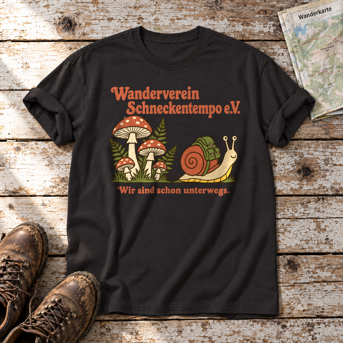 Wanderverein Schneckentempo – T-Shirt Unisex in Schwarz, flach liegend auf alten Holzbrettern mit Wanderschuhen und Wanderkarte.