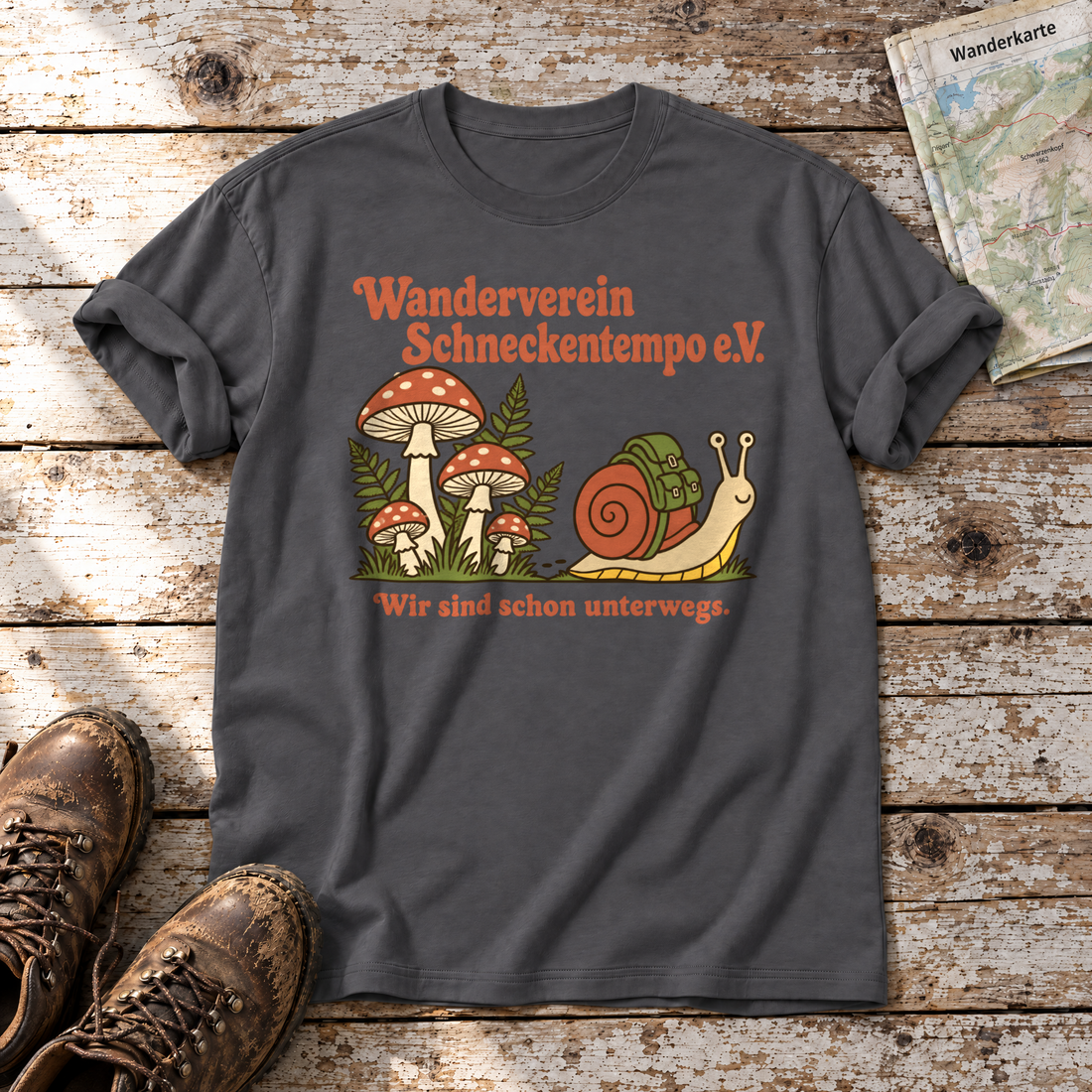 Wanderverein Schneckentempo – T-Shirt Unisex in Dunkelgrau, flach liegend auf alten Holzbrettern mit Wanderschuhen und Wanderkarte.