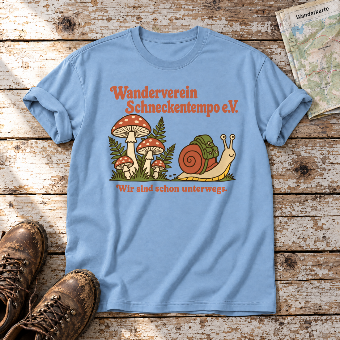 Wanderverein Schneckentempo – T-Shirt Unisex in Himmelblau, flach liegend auf alten Holzbrettern mit Wanderschuhen und Wanderkarte.